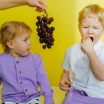 A boy enjoying grapes while sitting next to his curious sister indoors against a yellow background.